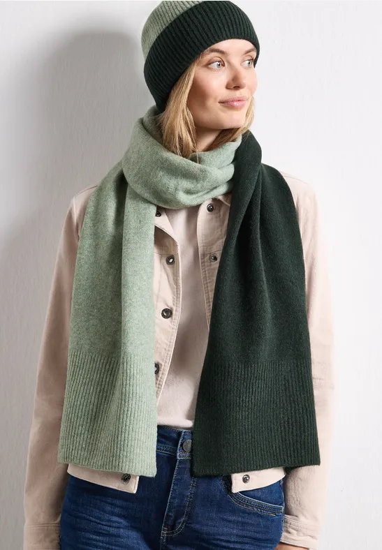 Frau mit grüner Strickmütze und zweifarbigem Schal trägt helle Jacke und Jeans, Blick zur Seite.