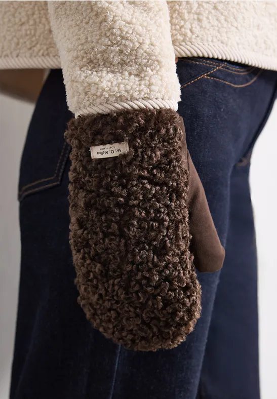 Dunkelbrauner, flauschiger Teddyfell-Fäustling vor Jeans und cremefarbener Teddyjacke.