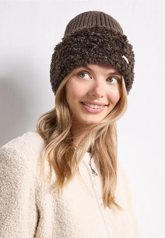 Junge Frau lächelt, trägt beige Teddyjacke und braune, flauschige Strickmütze.