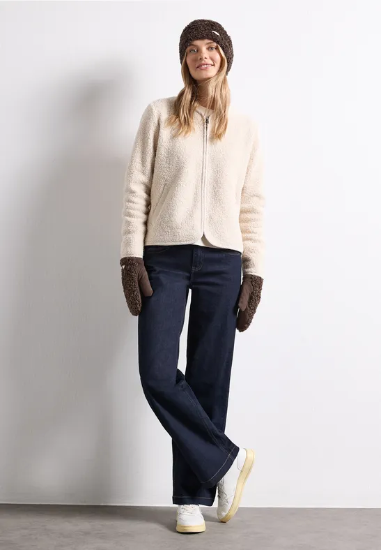 Eine junge Frau in heller Strickjacke, dunkler Jeans, braunen Fäustlingen und Mütze posiert lächelnd.