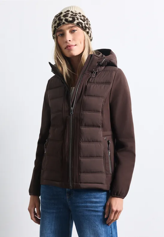 Frau in brauner Steppjacke, Jeans und Leopardenmütze vor hellem Hintergrund.