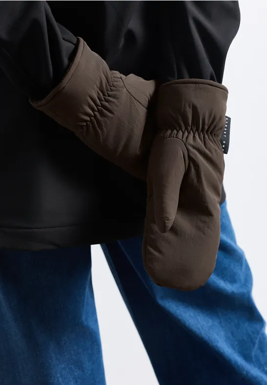 Person trägt schwarze Jacke, blaue Jeans und braune Fäustlinge, Nahaufnahme vor neutralem Hintergrund.