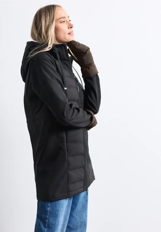 Frau im Profil trägt schwarze Jacke mit Kapuze, braune Handschuhe und blaue Jeans, vor hellem Hintergrund.