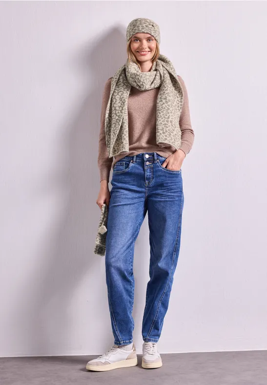 Frau im lässigen Look mit beiger Strickmütze und Schal, braunem Pullover, blauen Jeans und weißen Sneakers.