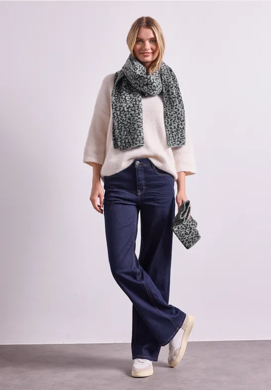Vrouw in crème trui, blauwe jeans, beige sneakers met groene gebreide sjaal en bijpassend tasje.