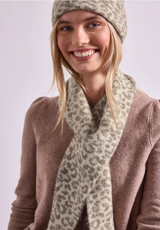 Lächelnde Frau mit gestrickter Mütze und Leopardenmuster-Schal, trägt braunen Pullover.