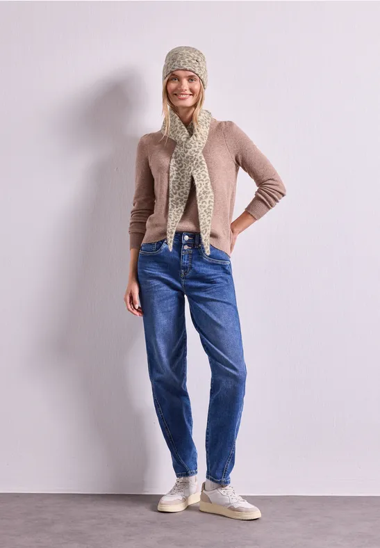 Frau in Beanie, Schal, beige Strickjacke, blauen Jeans und Sneakern steht lächelnd vor heller Wand.