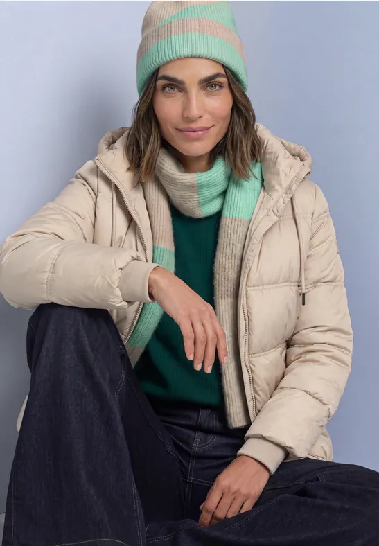 Femme portant une doudoune beige, bonnet et écharpe verts et beiges, pull vert et pantalon foncé, souriante.