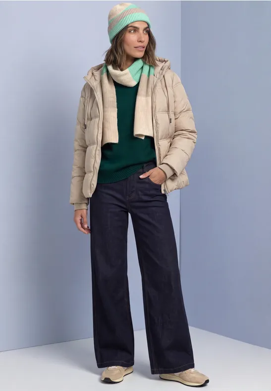 Femme en studio bleu clair portant une doudoune beige, pull vert, jean large foncé, bottes beige et bonnet pastel