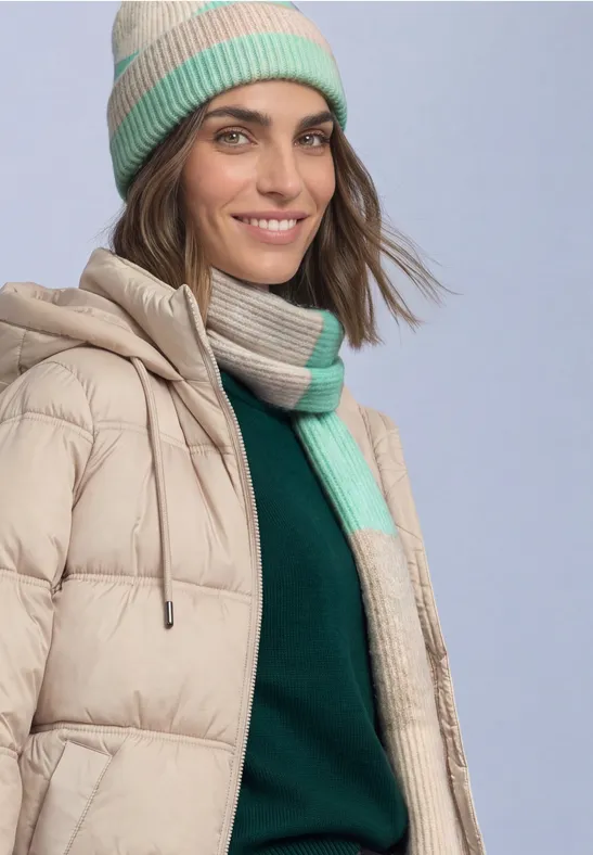 Une femme souriante porte une doudoune beige, un pull vert, un bonnet et une écharpe beige-vert.