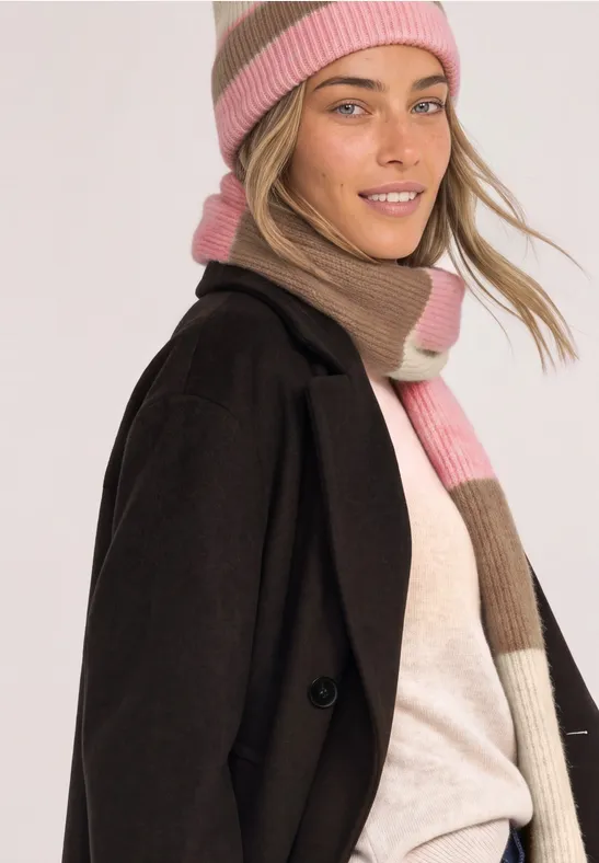 Junge Frau in schwarzem Mantel und rosa-beigem Strickschal und Mütze lächelt leicht.