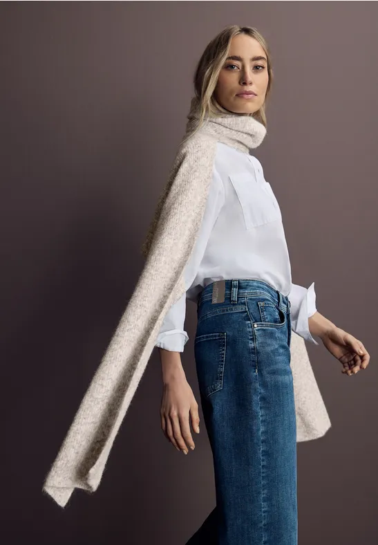 Frau mit weißer Bluse, blauen Jeans und langem beigem Schal vor grauem Hintergrund.