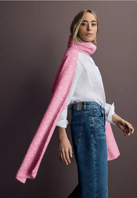 Femme en chemise blanche, jean et longue écharpe rose debout devant un fond uni.