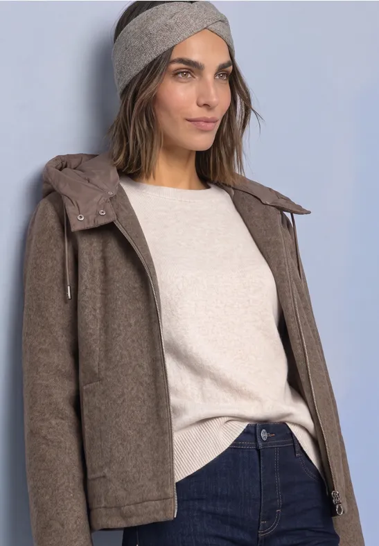 Frau in brauner Jacke, beigem Pullover, Jeans und Stirnband vor hellem Hintergrund.