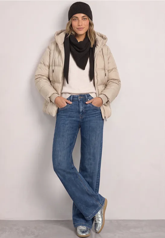 Femme en doudoune beige, bonnet et écharpe noirs, pull blanc, jean bleu et baskets argentées.