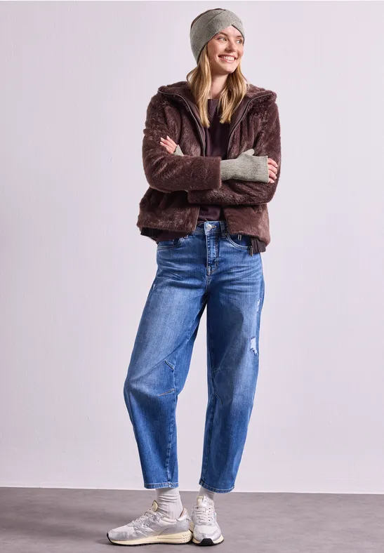 Eine lächelnde Frau in Wintermode mit brauner Teddyjacke, Mütze, Jeans und weißen Sneakern.