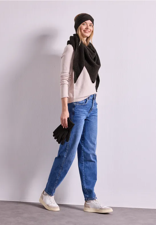 Junge Frau in Jeans, Pullover, großem schwarzem Schal und Mütze hält Handschuhe und lächelt.