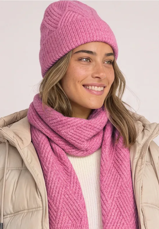 Frau mit pinker Strickmütze und passendem Schal, beiger Daunenjacke und freundlichem Lächeln.