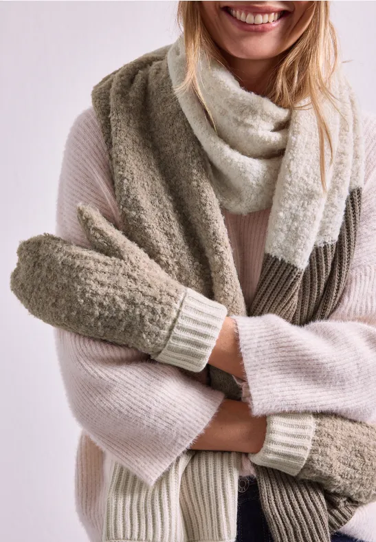 Frau in hellem Pullover mit zweifarbiger Strickschal und weichen Fäustlingen lächelt.