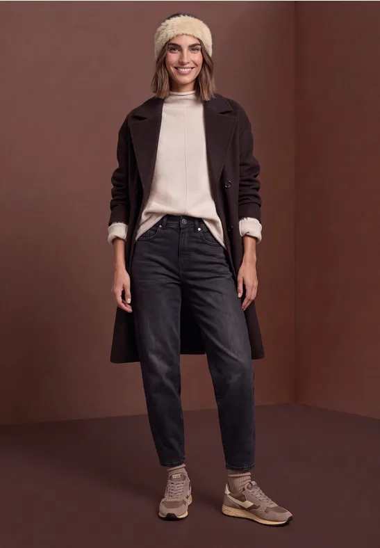 Frau in dunkelbrainem Mantel, schwarzer Jeans und beigen Accessoires vor braunem Hintergrund.