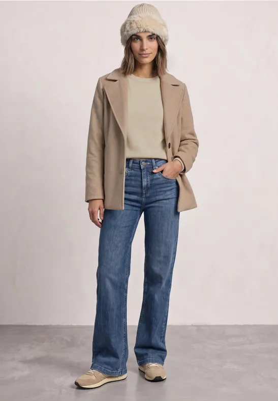 Vrouw in crème trui, beige jas, blauwe bootcut jeans en een gebreide muts.
