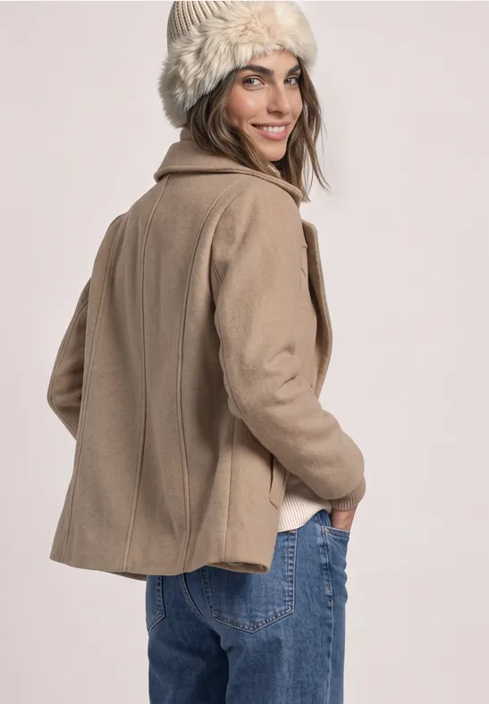 Een vrouw in een beige jas, blauwe jeans en een witte gebreide muts kijkt over haar schouder.