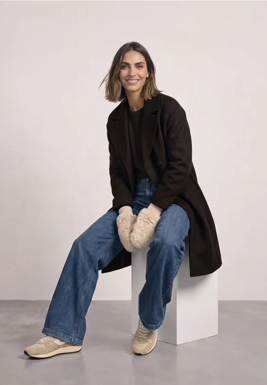 Eine lächelnde Frau sitzt auf einem weißen Block, trägt Jeans, Mantel, Fellhandschuhe und Sneaker.