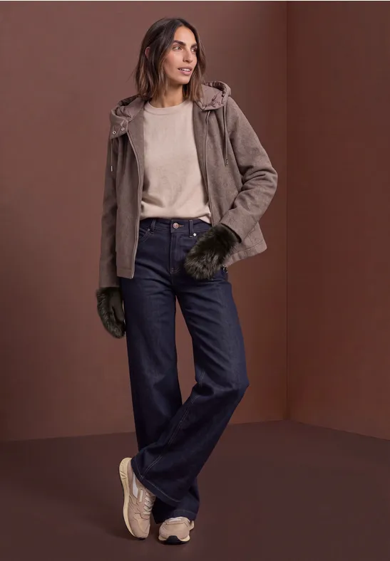 Frau in brauner Jacke, hellem Pullover, weiten Jeans und Handschuhen vor braunem Hintergrund.