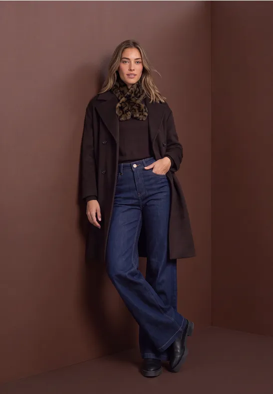 Eine Frau lehnt an brauner Wand, trägt Mantel, Rüschenbluse und weite Jeans.