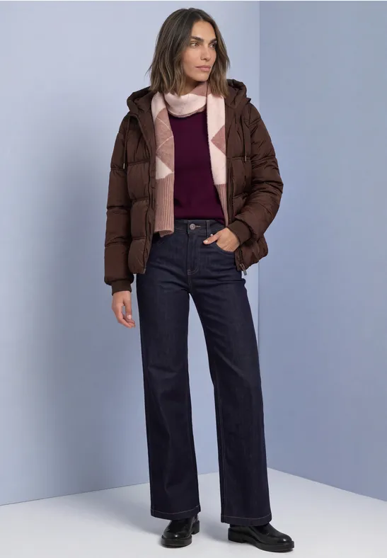 Frau in brauner Steppjacke, Bordeaux-Pullover, Schal und dunklen Jeans vor neutralem Hintergrund.