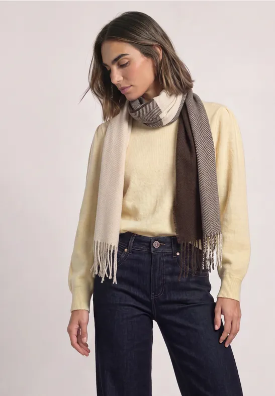 Frau in gelblichem Pullover, dunkler Jeans und zweifarbigem Schal, steht vor neutralem Hintergrund.