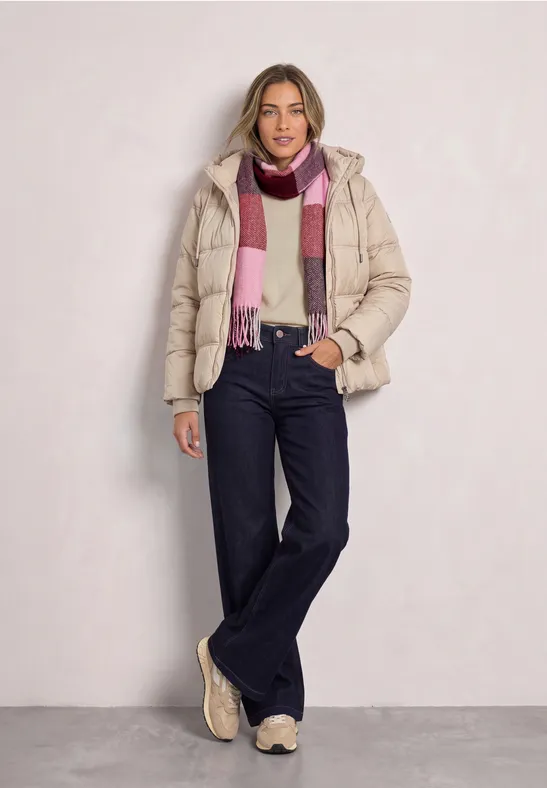 Frau trägt beige Steppjacke, Jeans, Schal und Sneakers in legerem Winterlook vor hellem Hintergrund.