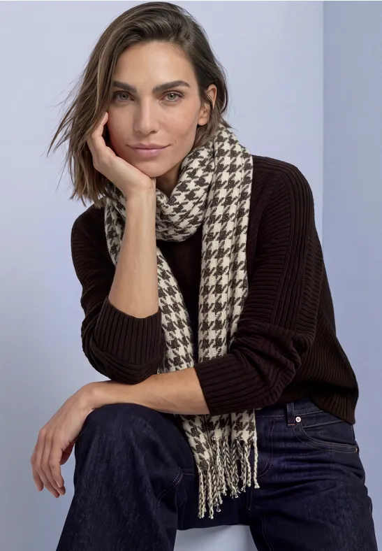 Eine Frau in dunklem Pullover und Jeans mit Hahnentritt-Schal sitzt lässig, Hand an der Wange.