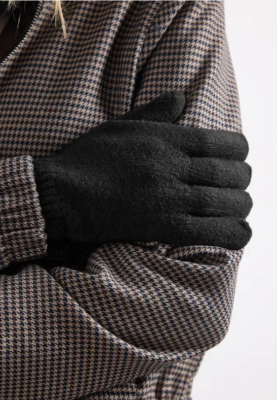 Close-up: persoon in pied-de-poule jas met zwarte handschoenen, armen gekruist voor de borst.
