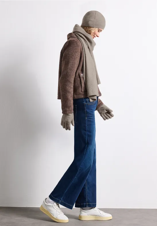 Frau in Winterkleidung mit Mütze, Schal, Handschuhen, brauner Jacke, blauen Jeans und weißen Sneakers.