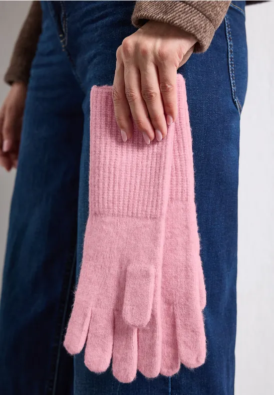 Eine Person in Jeans hält ein Paar rosa Strickhandschuhe in der Hand.