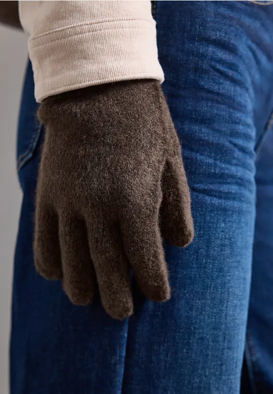 Nahaufnahme einer Hand mit flauschigem dunkelbraunen Handschuh vor blauer Jeans und hellem Oberteil.