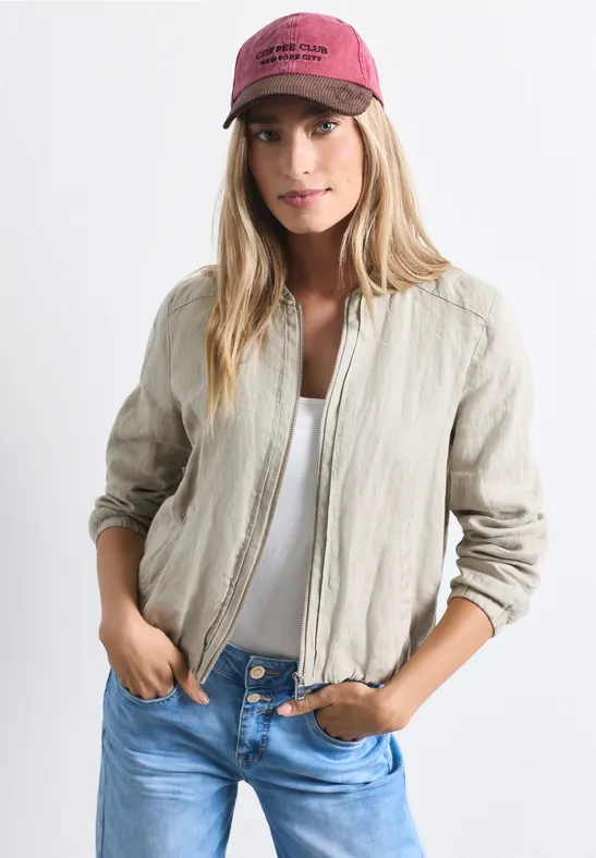 Junge Frau in heller Leinen-Blousonjacke, weißem Top, blauer Jeans und roter Kappe posiert lässig.
