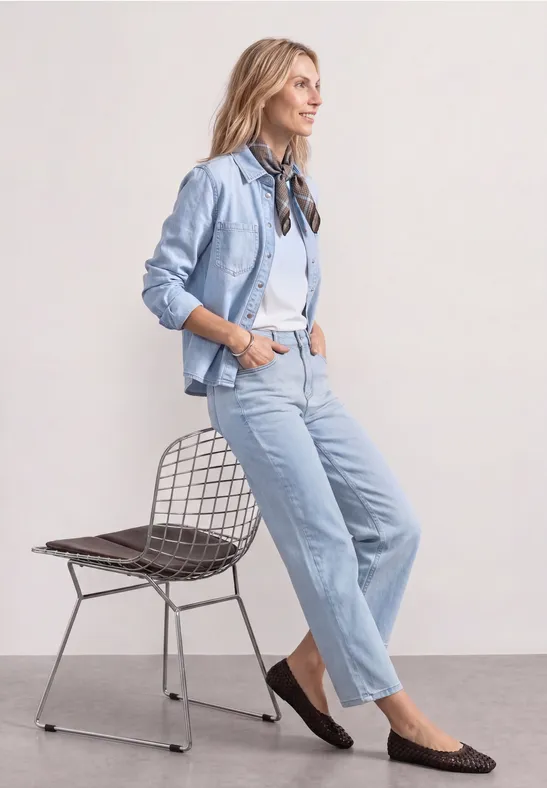 Eine junge Frau in hellem Jeansoutfit lehnt lässig an einem Drahtstuhl.
