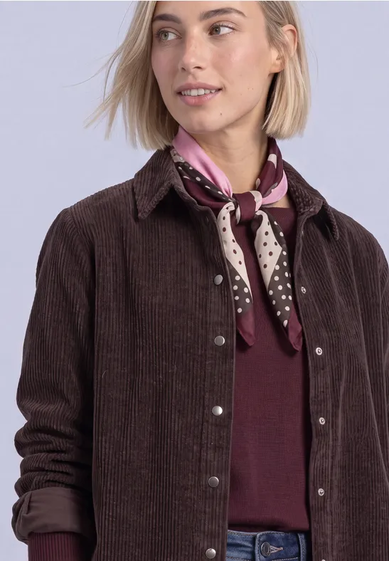 Frau mit blondem Bob trägt braune Cordjacke, weinrotes Shirt und gepunktetes lila Halstuch, schaut seitlich.