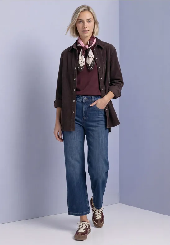 Frau in lockerem Casual-Outfit: dunkelrote Bluse, dunkelbraune Hemdjacke, Jeans, Halstuch und Boots.