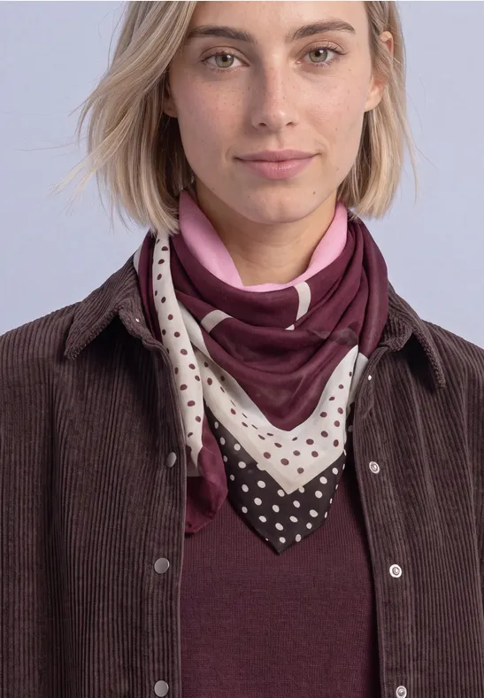 Frau mit blondem Bob trägt braune Cordjacke, weinrotes Shirt und gemustertes Halstuch vor hellem Hintergrund.