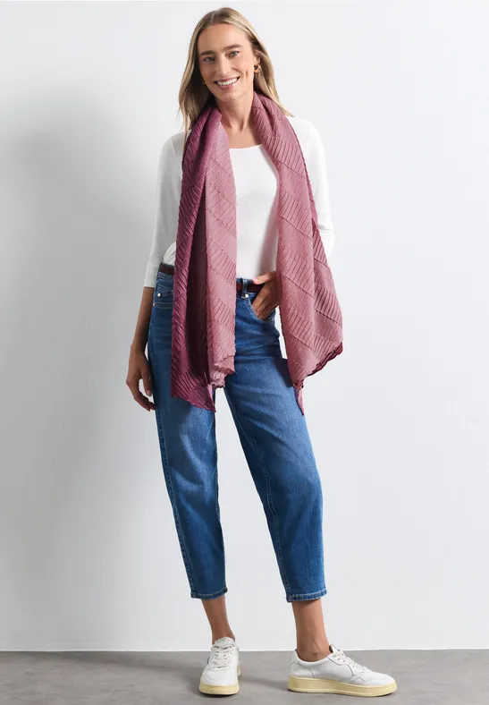 Frau lächelnd in weißem Shirt, blauen Jeans und rosa Schal vor weißem Hintergrund.