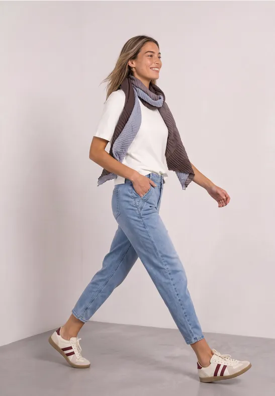 Frau spaziert fröhlich in Jeans, weißem T-Shirt, Sneakern und Schal vor schlichtem Hintergrund.