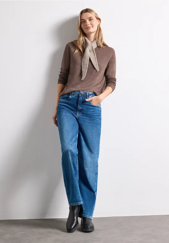 Vrouw voor witte muur, draagt bruine trui, sjaaltje, wijde blauwe jeans en zwarte laarsjes.