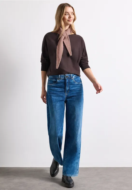 Frau in braunem Pullover, Halstuch, weiten Jeans und schwarzen Schuhen vor weißem Hintergrund.