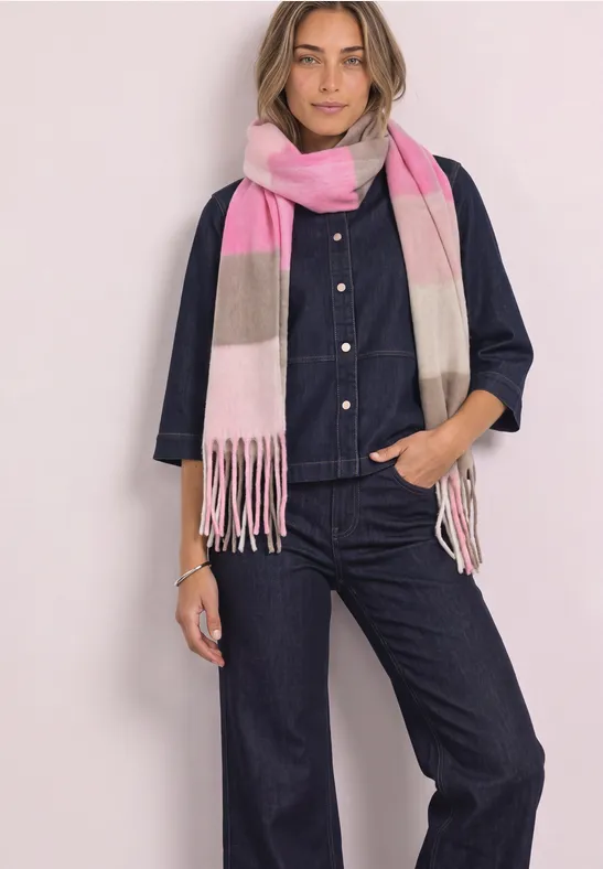 Eine Frau trägt einen dunkelblauen Denim-Overall und einen rosa-beigen Schal, sie steht vor hellem Hintergrund.
