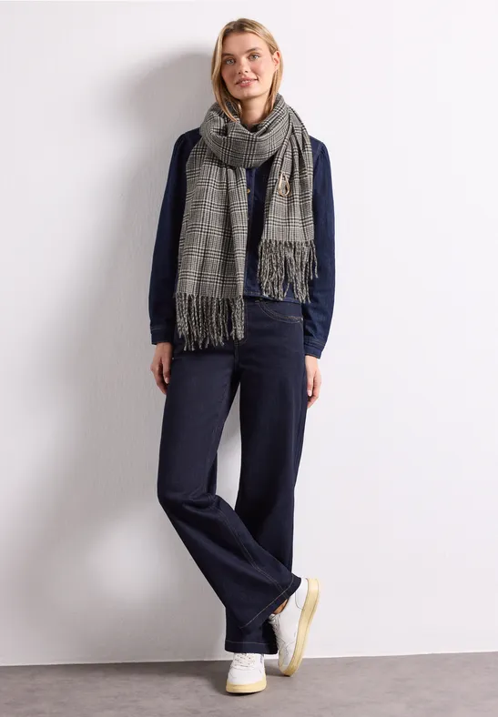 Frau in lässigem Outfit: dunkelblaue Jeans, Pulli, grauer Schal und weiße Sneaker vor weißem Hintergrund.