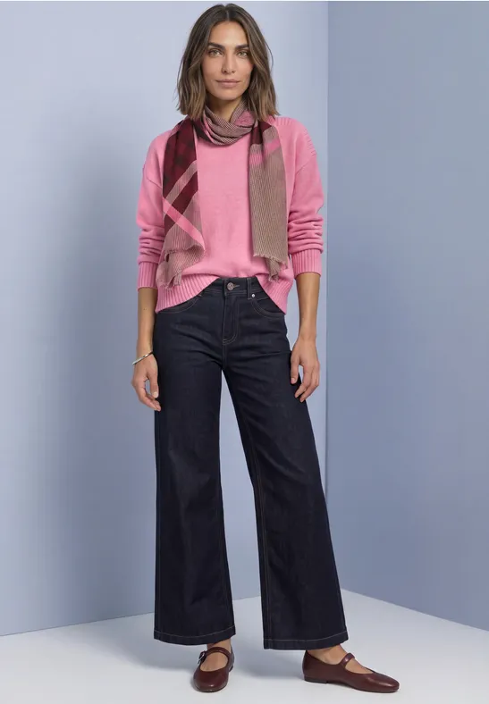 Eine Frau trägt pinken Pullover, Schal, weite Jeans und Loafer, steht vor neutralem Hintergrund.