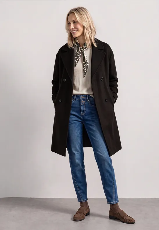Frau mit schwarzem Mantel, blauer Jeans, weißer Bluse und Halstuch, dazu braune Schuhe.